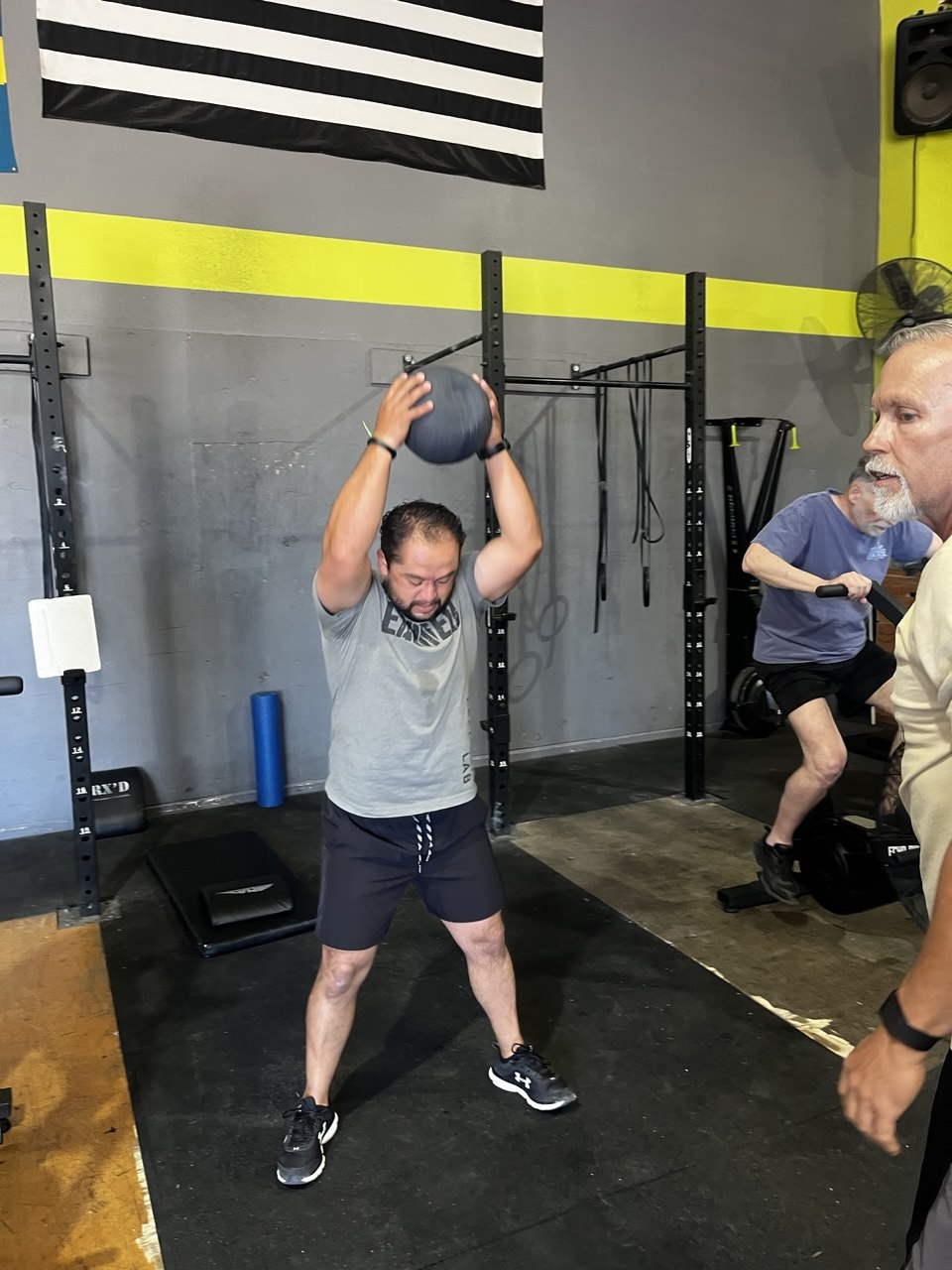 Member pressing a medicine ball overhead with a rower behind