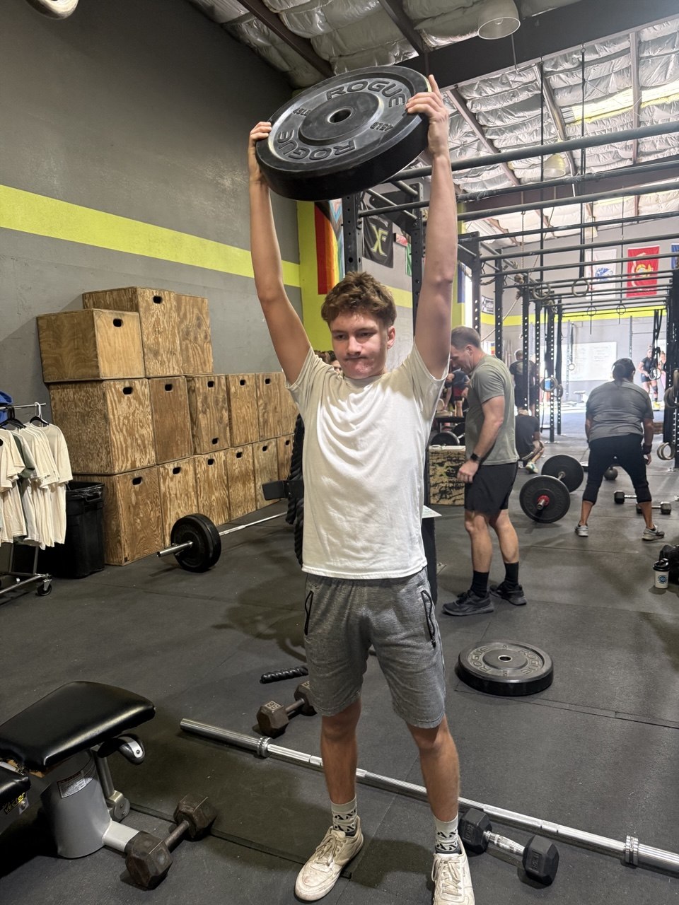Member holding a bumper plate overhead on a busy gym floor
